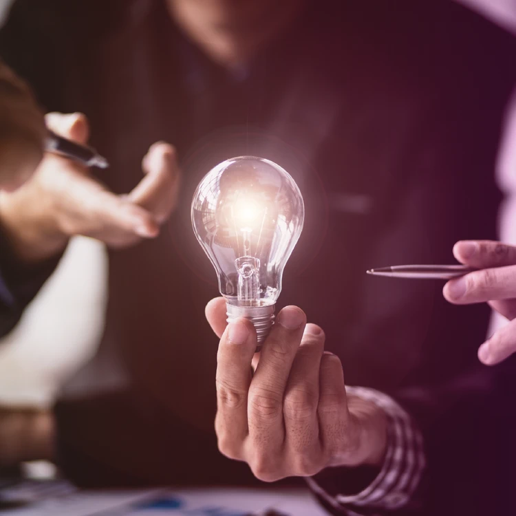 Hands of colleagues in a meeting presenting a lit lightbulb, symbolizing business innovation.