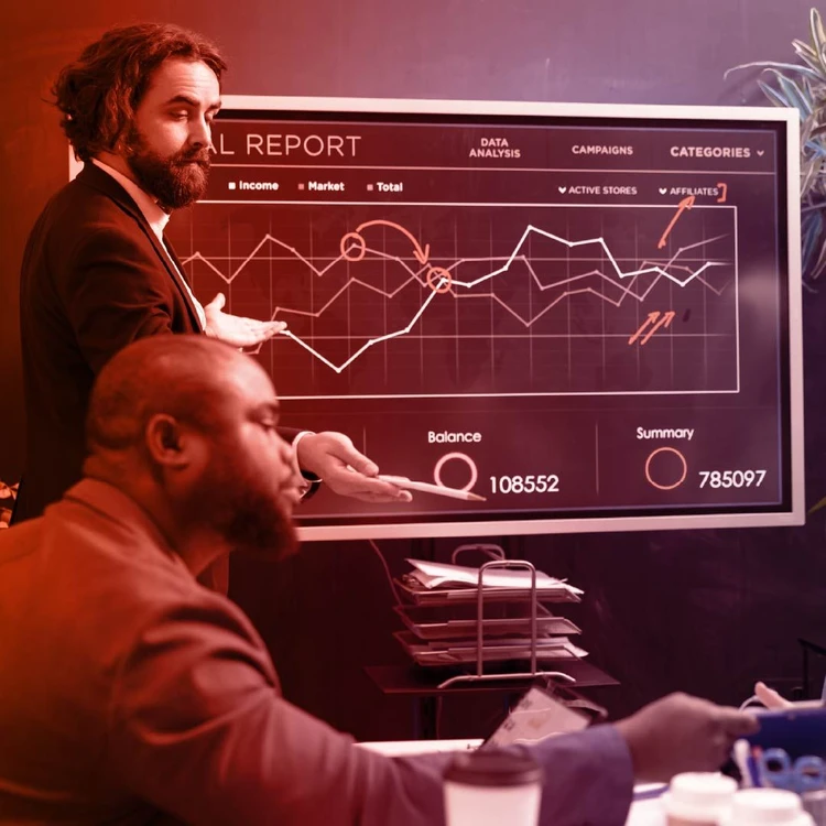 Two professionals, one standing and gesturing towards a large screen displaying analytical charts, and another sitting at a desk, discuss an annual report in an office.