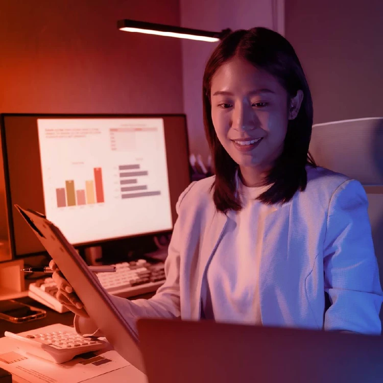 A professional observes a monitor while holding a folder of documents. Behind her is a second computer displaying bar charts.