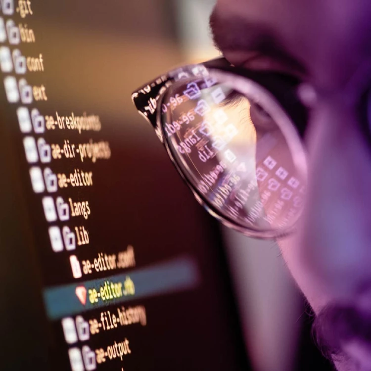 The eyeglasses of a male profile reflect strings of code from a monitor.