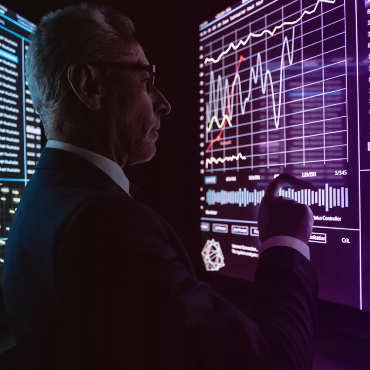 A man illuminated by the light from the monitors observes and points to a line graph on a large touch screen displaying data and diagrams.